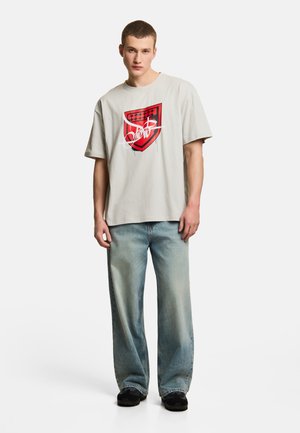 Młody mężczyzna w luźnej jasnoszarej koszulce z czerwoną i czarną grafiką oraz wyblakłych szerokich niebieskich dżinsach, stojący na białym tle.