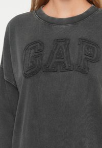Tmavě šedá mikina s vyvýšeným černým vyšitým nápisem "GAP" na hrudi, s vroubkovaným výstřihem a volným střihem. Měkká, strukturovaná tkanina.