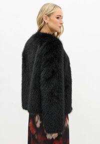 Chaqueta de piel sintética negra con un diseño recortado y superficie texturizada. Presenta mangas largas y un cuello alto, combinada con un vestido floral en la parte inferior.