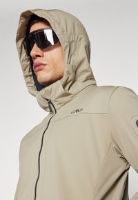Beige hooded jacket with a front zipper and CMP logo. Features smooth fabric, a snug fit, and a contrasting black interior lining.