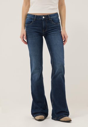 Donna che indossa jeans bootcut blu scuro e scarpe beige, in piedi con le braccia rilassate davanti a uno sfondo chiaro e semplice.