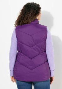 Lila Puffer-Weste mit Zickzack-Steppung, hohem Kragen und glatter Textur, kombiniert mit einem helllila Langarmshirt und Jeans.