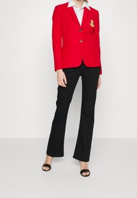 Röd blazer med guldknappar och broderade detaljer, tillsammans med svarta utsvängda byxor. Modellen bär svarta öppna skor, med en vit skjorta med krage.