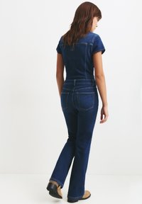 Granatowy dżinsowy kombinezon z krótkim rękawem, dopasowaną górą, rozszerzanymi nogawkami i tylnymi kieszeniami. Kontrastowe szycie podkreśla detale projektu.