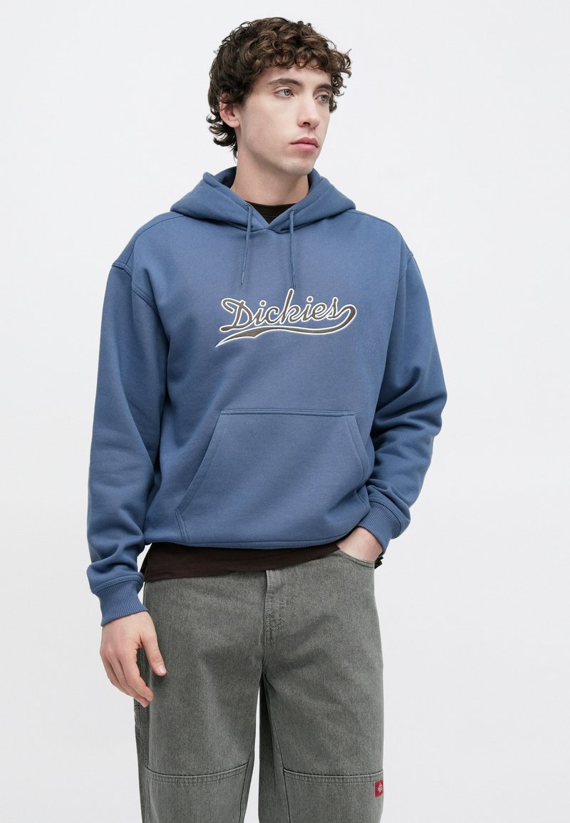 Felpa con cappuccio in cotone blu con tasca frontale e logo "Dickies" ricamato. Presenta un cappuccio con coulisse e polsini a costine. Indossata sopra pantaloni cargo grigi.