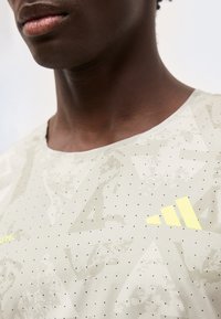 Κοντινό πλάνο ενός ατόμου που φοράει μια μπεζ μπλούζα Adidas με κίτρινο λογότυπο στο στήθος και μικρές μαύρες κουκκίδες σε όλο το ύφασμα.