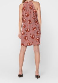 Vrouw in een mouwloos, knie-lange, stoffige roze jurk met een rood bloemenpatroon, staand met haar rug naar de camera, in zwarte kittenheels.