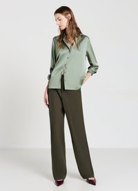 Grünes Satinhemd mit langen Ärmeln und Knöpfen, kombiniert mit hoch taillierten, weit geschnittenen dunkelgrünen Hosen und bordeauxfarbenen Absätzen.