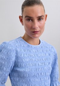 Woman with slicked-back hair wearing small hoop earrings and a light blue textured long-sleeve top against a plain background.