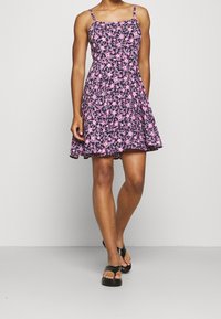 Blumenkleid in dunklem Lila mit pinken Blumen, tailliertem Oberteil, A-Linien-Rock und dünnen verstellbaren Trägern. Kombiniert mit schwarzen Plateausandalen.