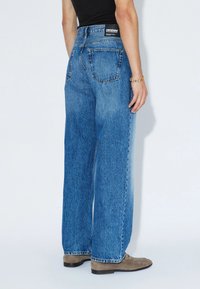 High-waisted blue denim jeans with wide legs, featuring a classic cut, back pockets, and a brand label at the waist. Fabric has a textured finish.