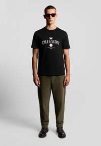 Camiseta de algodón negra con diseño de logo en blanco y rosa, combinada con pantalones verde oliva. Modelo con gafas de sol oscuras y botas negras.