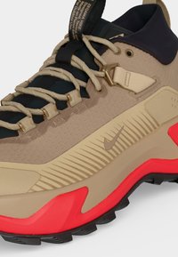 Μπεζ και μαύρο πεζοπορικό παπούτσι Nike με κόκκινες λεπτομέρειες στη σόλα, κορδόνια σχοινιού, υφασμένα πλαϊνά πάνελ και ετικέτα με την επιγραφή "Designed Tested Made On Planet Earth."