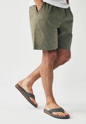 Mann trägt olivgrüne Shorts und graue Flip-Flops, steht mit den Händen in den Taschen vor weißem Hintergrund.
