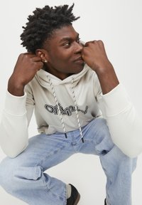 Sudadera gris claro con letras negras, cordones ajustables en blanco y negro; combinada con jeans azul claro y zapatillas oscuras.