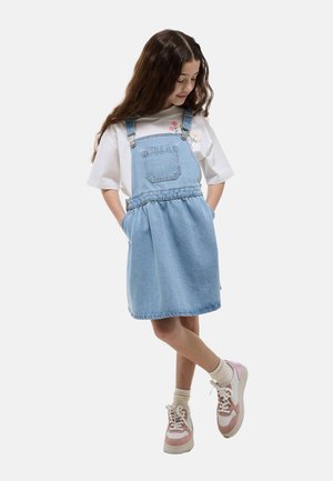 Jeune fille aux longs cheveux bruns portant un t-shirt blanc à fleurs, une robe salopette en denim bleu clair, des chaussettes crème et des baskets roses, debout les mains dans les poches.