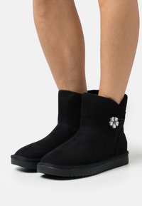 Stivali alla caviglia in suede nera con punta rotonda, dettagli cuciti, fodera soffice e un accento decorativo a forma di fiore sul lato. Suola piatta in gomma.