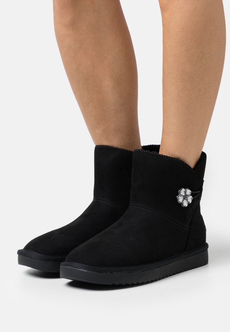 Stivali alla caviglia in suede nera con punta rotonda, dettagli cuciti, fodera soffice e un accento decorativo a forma di fiore sul lato. Suola piatta in gomma.