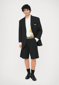 Črn blazer preko bele srajce z zlatimi vzorci, skupaj z črnimi kratkimi hlačami in črnimi supergami. Dodatki vključujejo blagovno znamko na pasu in podrobnosti na manšetah.