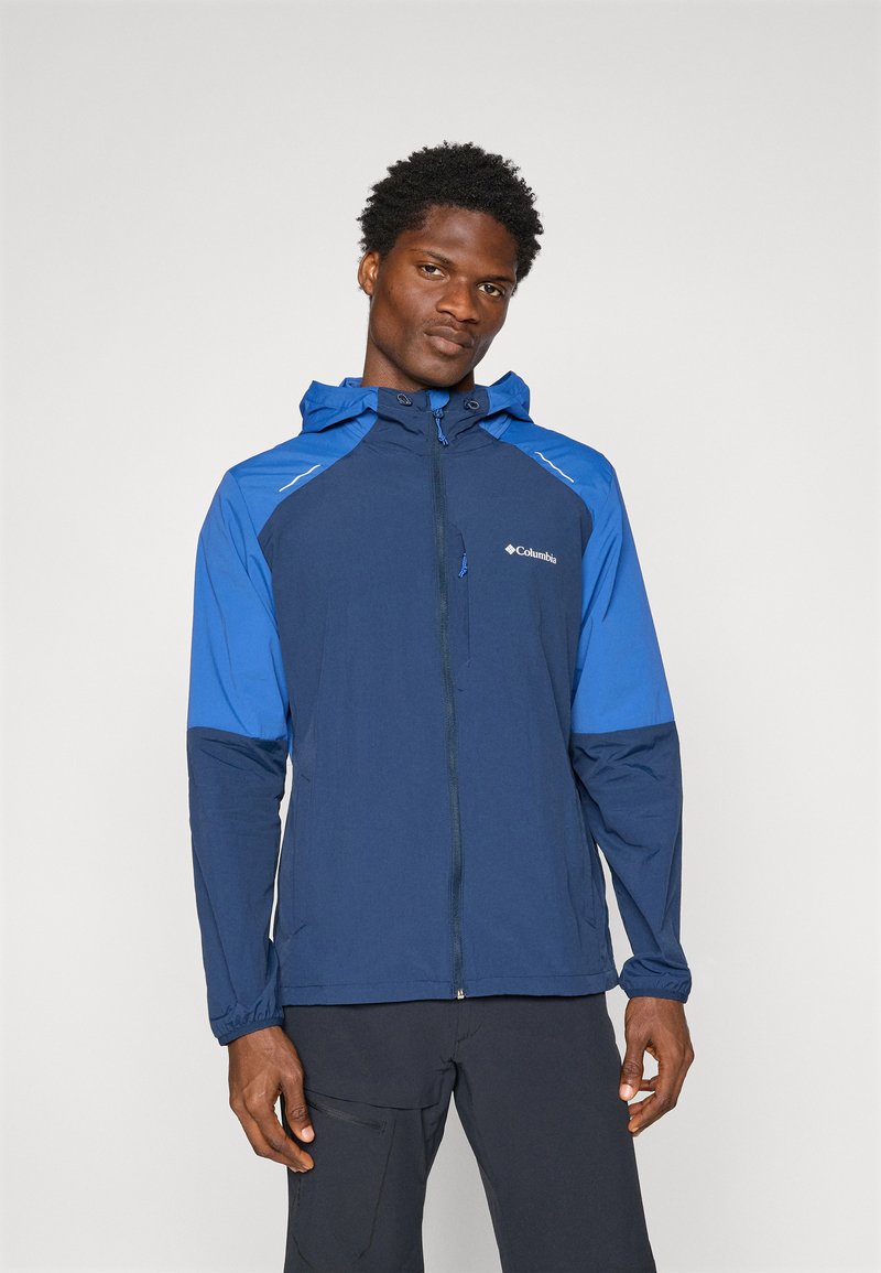 Veste résistante à l'eau avec un corps bleu marine et des manches bleues, dotée d'une capuche et d'une fermeture éclair à l'avant. Des détails réfléchissants sont visibles le long des coutures.