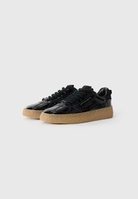 Baskets en cuir verni noir avec une épaisse semelle texturée beige et de grosses lacets noirs sur fond blanc.
