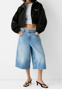 Svart kort kappa med elastiska manschetter, vit åtsittande skjorta, ljusblå vida denimculotter och svarta remsandaler med klack.