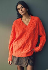 Helles orange gestricktes Sweatshirt mit V-Ausschnitt, das ein Rautenmuster und gerippte Bündchen aufweist. Kombiniert mit einem braunen Leder-Minirock.