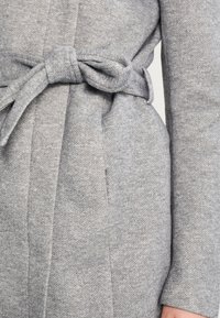 Abrigo gris texturizado con un amplio cinturón atado a la cintura, que presenta una silueta limpia y un diseño minimalista.