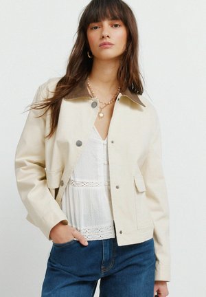 Frau mit langen braunen Haaren trägt eine cremefarbene Jacke mit braunem Kragen über einer weißen Spitzenbluse und blauen Jeans, steht vor einem schlichten Hintergrund.