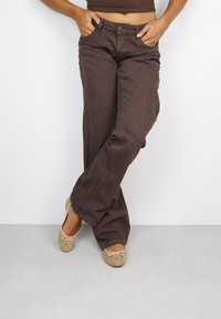 Pantalones marrones de pierna ancha, hechos de algodón suave con una textura lisa. Presentan bolsillos delanteros y un cierre clásico de botón. Combinados con zapatos planos beige.