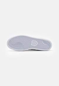 Semelle de chaussure avec une semelle extérieure en caoutchouc blanc texturé, présentant un motif en chevrons et un emblème central dans le design.