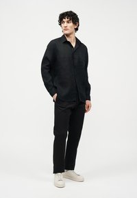 Hombre con cabello rizado, vestido con camisa negra de manga larga, pantalones negros y zapatillas blancas, de pie con una mano en el bolsillo, sobre fondo blanco.