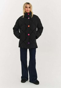 Czarna pikowana kurtka puchowa z wysokim kołnierzem, ozdobiona dużymi, kolorowymi guzikami w kolorach różowym, pomarańczowym i fioletowym. Noszona na dzwonowatych dżinsach.