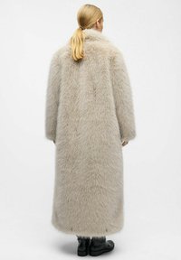 Frau mit blonden Pferdeschwanz, die einen langen beigen Kunstpelzmantel und schwarze Knöchelstiefel trägt, steht vor einem schlichten weißen Hintergrund.