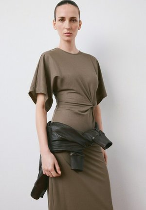 Ženska z lasmi, povezanimi nazaj, oblečena v rjavo obleko z vozličasto podrobnostjo na pasu in s črno usnjeno jakno, zavezano okoli bokov, ob belem zidu.