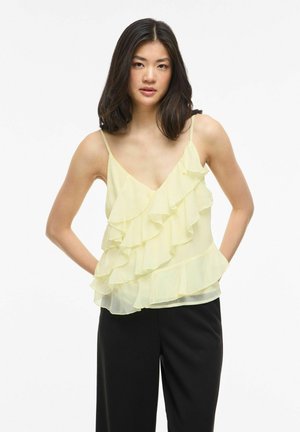 Ung kvinne med skulderlangt svart hår iført en blekgul topp med volanger uten ermer og svarte bukser, hendene i lommene, nøytral bakgrunn.