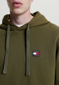 Olivgrüner Hoodie mit einer Fronttasche, Kordeln und einem kleinen rechteckigen „TOMMY JEANS“ Logo-Patch in Marineblau, Rot und Weiß. Weicher Stoff.