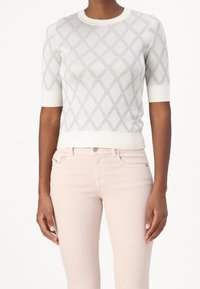 Mujer con un suéter blanco de manga corta con un patrón de diamantes gris y unos pantalones de mezclilla de tiro alto en rosa claro, de pie frente a un fondo blanco.