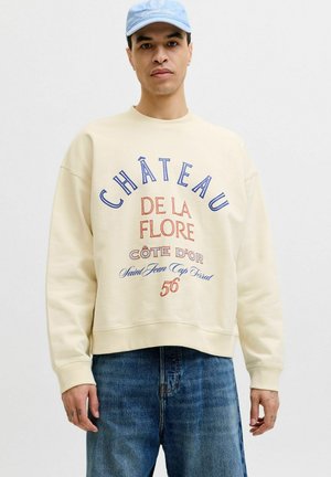 Mann trägt hellbeige Sweatshirt mit dem Schriftzug „Château de la Flore Côte d'Or 56“ und blaue Baseballkappe, steht vor einfarbigem Hintergrund.
