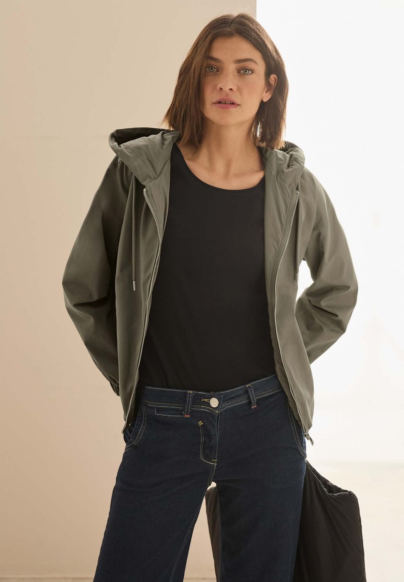 Lehká olivově zelená bunda s kapucí, zipovým uzávěrem a šňůrkami. Spárovaná s černým tričkem a tmavě modrými džíny, ukazující ležérní vzhled.