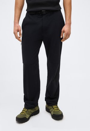 Mand iført sorte udendørs bukser med indbygget bælte og olivengrønne vandresko med gule snørebånd, stående på en hvid baggrund.