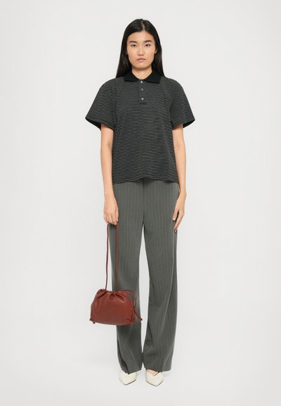 Femeie îmbrăcată cu un polo cu dungi negre și albe, pantaloni largi gri cu dungi, încălțăminte albă cu vârful ascuțit, ținând o geantă de umăr maro cu șnur.