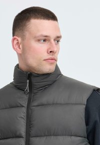Gepolsterte Weste aus dunkelgrauem Nylon mit hohem Kragen und Frontreißverschluss. Verfügt über horizontale Nähte und leichte Polsterung für Wärmeisolierung.