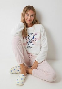 Bílá, huňatá mikina se grafikou Snoopy; růžové, vzorované pyžamové kalhoty; bílé pantofle s modrými srdíčky. Měkké materiály a volný střih.