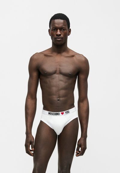 Αθλητικός άνδρας με σκούρο δέρμα που φοράει λευκά εσώρουχα Moschino με σύμβολο καρδιάς, στέκεται μπροστά σε απλό ανοιχτό φόντο.