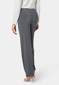 Grijze, wijde broek met een elastische tailleband en een soepele stof. Het model draagt naaktkleurige hoge hakken. De achterkant toont de pasvorm van het kledingstuk.