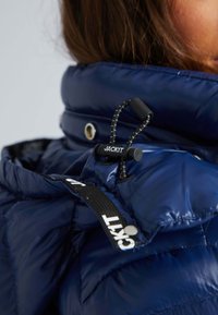 Gros plan sur le col d'une veste matelassée bleue avec un verrou de cordon réglable noir portant l'inscription "JACK1T" et une partie des cheveux bruns d'une personne visible.