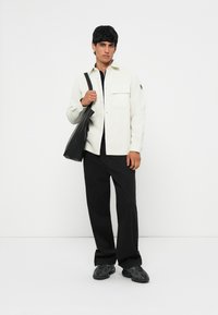 Camicia a maniche lunghe di un beige chiaro con texture, dotata di taschino sul petto, abbinata a pantaloni a gamba larga neri e sneakers scure, mentre tiene una borsa tote nera.