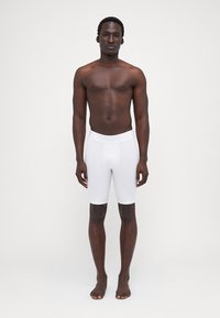 Hvide bokserumm shorts lavet af strækstof. Glat overflade, tætsiddende design, midt på låret længde. Modellen står barfodet mod en ensfarvet baggrund.