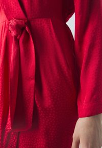 Gros plan d’une robe en tissu rouge texturé avec une ceinture nouée à la taille et la main droite d’une personne détendue sur le côté.